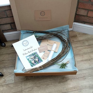 K&ouml;nnte beinhalten: Ein brauner Karton mit einer blauen und wei&szlig;en Seidenpapierauskleidung. Die Schachtel enth&auml;lt ein Kit zum Bau von Weidenvogelh&auml;uschen. Das Kit enth&auml;lt Weidenzweige, Anleitungen und ein kleines Holzschild. Das Schild sagt "Danke".