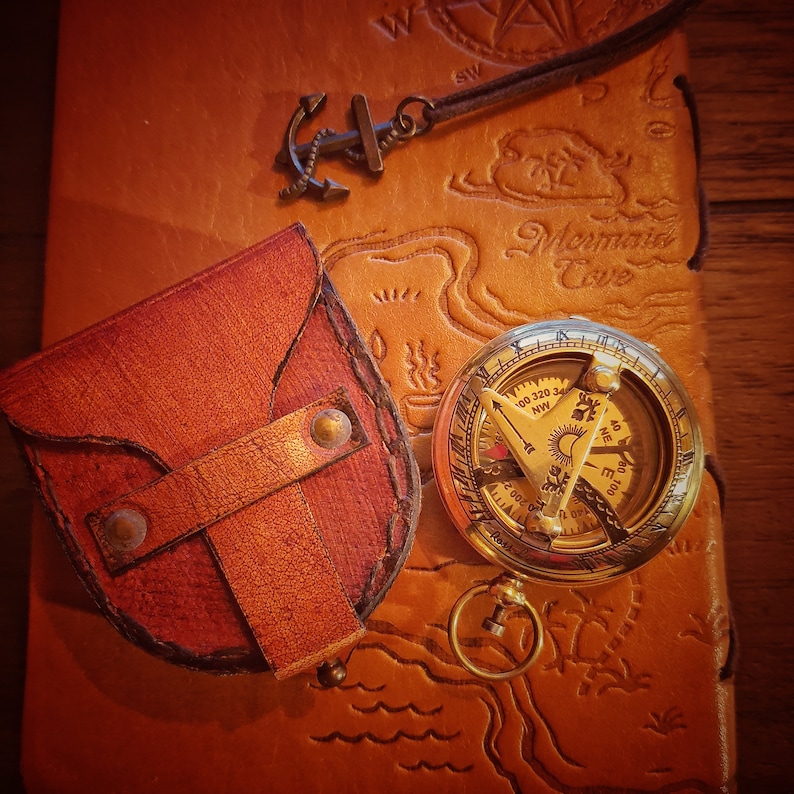 K&ouml;nnte beinhalten: Ein antiker Kompass aus Messing mit einem Lederetui und einem Anker-Anh&auml;nger. Der Kompass hat ein detailliertes Design mit Markierungen f&uuml;r Richtungen und einer Sonnenuhr. Das Lederetui ist braun und hat eine Klappe, die mit einem Druckknopf geschlossen wird.