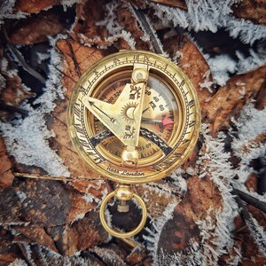 K&ouml;nnte beinhalten: Ein Kompass aus Messing mit einem Sonnenuhr und einer roten Nadel. Der Kompass liegt auf einem Hintergrund aus braunen Bl&auml;ttern und Frost.