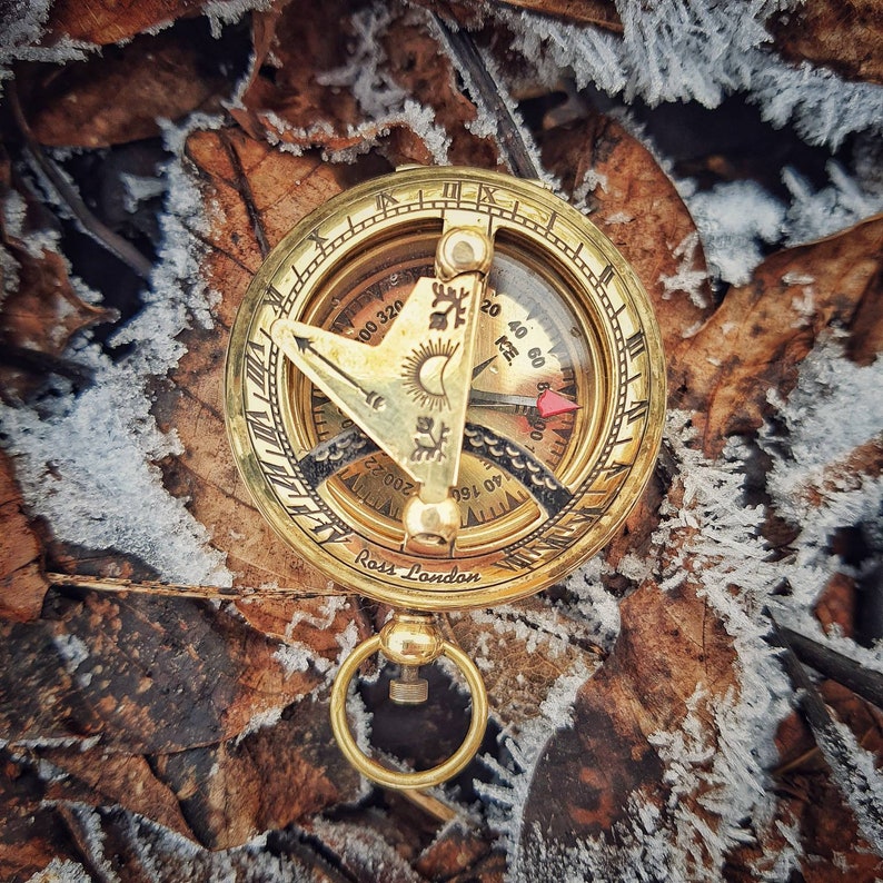 K&ouml;nnte beinhalten: Ein Kompass aus Messing mit einem Sonnenuhr und einer roten Nadel. Der Kompass liegt auf einem Hintergrund aus braunen Bl&auml;ttern und Frost.
