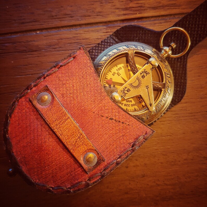K&ouml;nnte beinhalten: Ein antiker Kompass aus Messing mit einem Lederetui. Das Kompassgesicht hat r&ouml;mische Ziffern und Markierungen f&uuml;r Richtungen. Das Etui ist aus braunem Leder mit N&auml;hten an den R&auml;ndern.