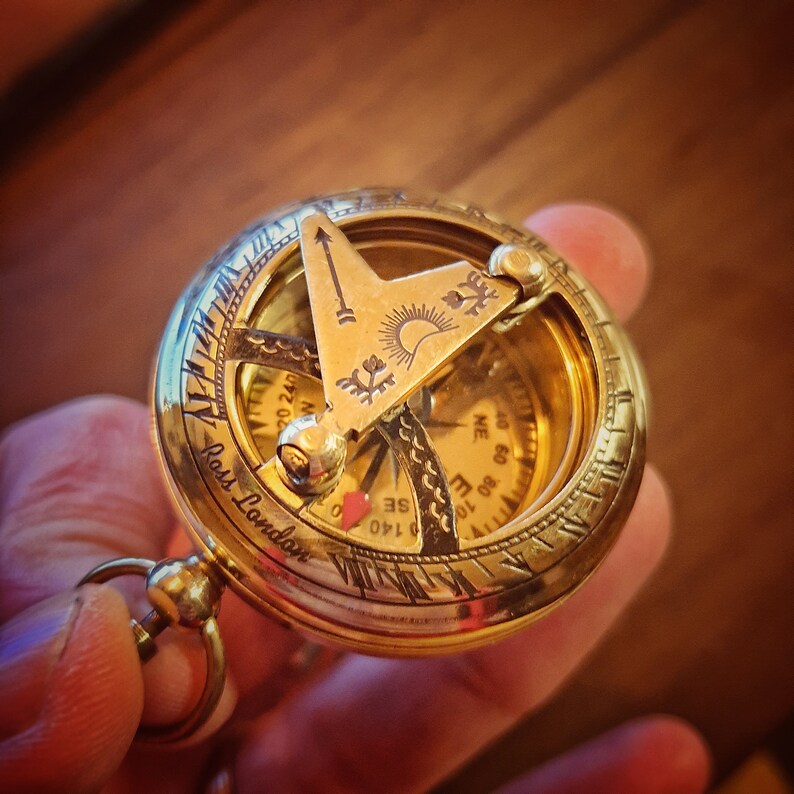 K&ouml;nnte beinhalten: Ein goldfarbener Kompass mit einer Kompassrose und einer Sonnenuhr. Der Kompass ist mit den Worten "Ross London" graviert.