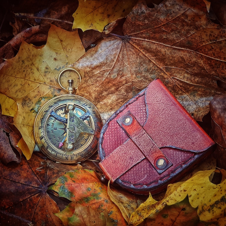 K&ouml;nnte beinhalten: Ein Kompass aus Messing mit einer braunen Ledertasche auf einem Bett aus Herbstlaub. Der Kompass hat ein goldenes Zifferblatt mit schwarzen Markierungen und eine Nadel, die nach Norden zeigt. Die Tasche hat eine Klappe mit einem Schnallenverschluss.