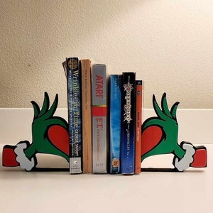 May include: Green bookends shaped like the Grinch's hands holding red hearts. The bookends are holding up a variety of books, including "Wrinkles in Time", "E.T. The Extraterrestrial", "Dragonlance Chronicles Vol. 3", and "Madvillainy".