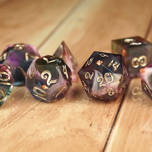 Könnte beinhalten: Sieben mehrseitige Würfel in verschiedenen Formen mit einem wirbelnden lila, blauen und rosa Design. Die Zahlen sind goldfarben. Geeignet für Tabletop-Spiele.