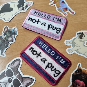 May include: Two embroidered patches with the text "Hello I'm not a pug" in white on a pink and blue background. The patches are surrounded by various dog stickers.