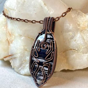 以下が含まれることがあります： 手作りの銅線巻きペンダントネックレス。ペンダントは楕円形の黒と白の石が特徴です。銅線は石の周りに複雑に織り込まれており、ネックレスは銅のチェーンが付いています。