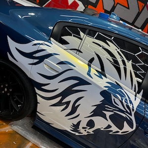 Puede incluir: Un coche azul con un gráfico blanco y negro de un tigre rugiente en el lateral. El gráfico del tigre está estilizado con llamas.