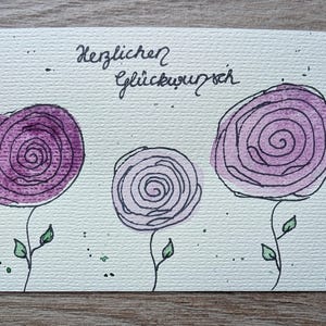 May include: A watercolour greeting card featuring three stylized purple roses. The card has the German phrase "Herzlichen Glückwunsch" written in black script. The roses have green leaves and stems against a textured white background.