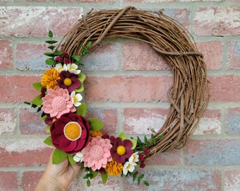 May include: A felt flower wreath with a grapevine base. The wreath is decorated with pink, red, and yellow felt flowers. The flowers are arranged in a cluster on the left side of the wreath.