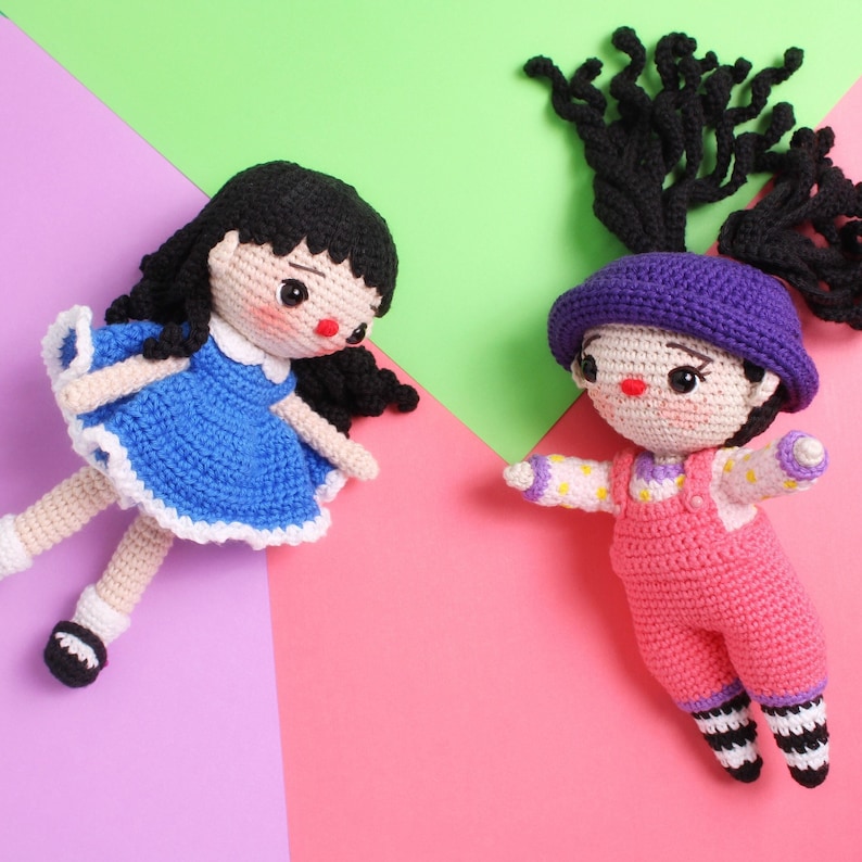 May include: Two crocheted dolls, one with a blue dress and black hair, the other with a pink dress, purple hat, and black hair.