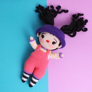 May include: A crocheted doll with black hair, a purple hat, and a pink and white outfit. The doll is wearing overalls and has black and white striped legs.