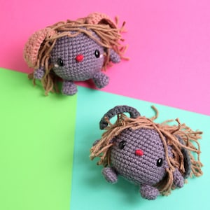 May include: Two crocheted gray plush toys with brown yarn hair and pink ears. The toys are sitting on a pink and green background.