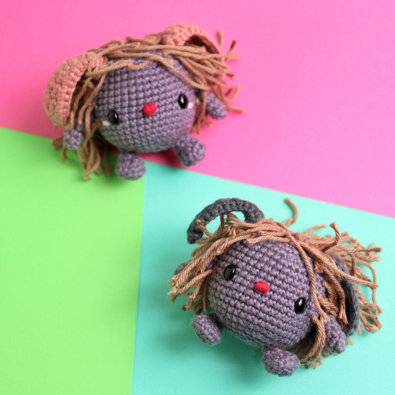 May include: Two crocheted gray plush toys with brown yarn hair and pink ears. The toys are sitting on a pink and green background.