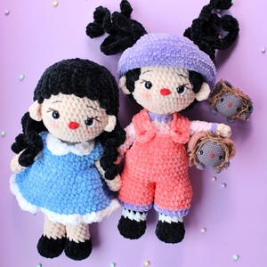 May include: Two handmade crochet dolls. One doll wears a blue dress with a white collar, black shoes, and black hair. The other doll wears pink overalls, a purple hat, and black shoes. Each doll has a small crochet animal.