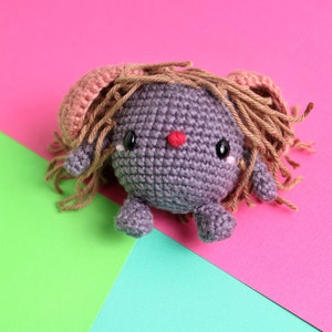 May include: A gray crocheted toy with a pink and brown yarn hairdo. The toy has large black eyes and a red nose. The toy is sitting on a pink, green, and blue background.