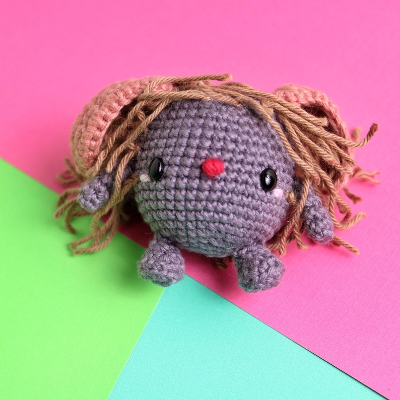 May include: A gray crocheted toy with a pink and brown yarn hairdo. The toy has large black eyes and a red nose. The toy is sitting on a pink, green, and blue background.