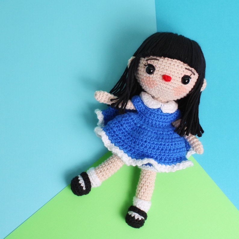 May include: A crocheted doll with black hair, a blue dress with white trim, and red cheeks. The doll is wearing black shoes and is posed on a green surface.