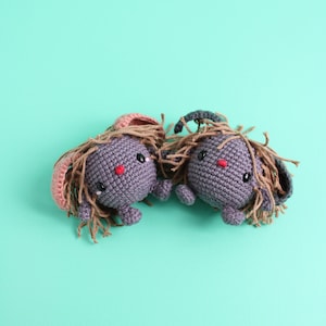 May include: Two crocheted gray bunny plushies with brown yarn hair, pink and black ears, and red noses. The plushies are facing each other on a turquoise background.