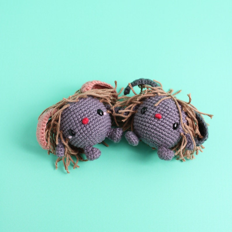 May include: Two crocheted gray bunny plushies with brown yarn hair, pink and black ears, and red noses. The plushies are facing each other on a turquoise background.