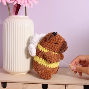 May include: A crocheted bee toy in brown and yellow with white wings. The plushie has a black spot and holds a white string. It's positioned near a cream vase with pink flowers. The bee's measurements are approximately 10 cm in length.