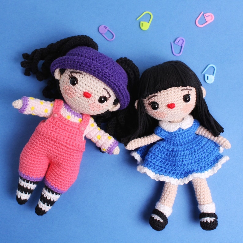 May include: Two crocheted dolls with black hair. One doll is wearing a pink and white dress with overalls. The other doll is wearing a blue dress with white trim. Both dolls have red yarn for their mouths.