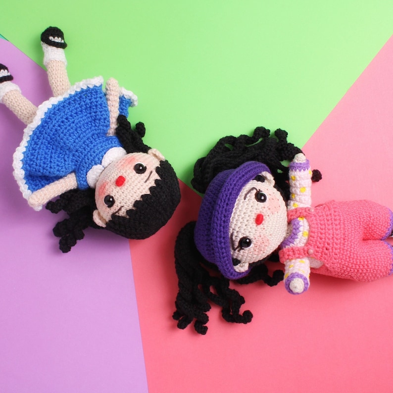 May include: Two crocheted dolls with black hair and smiling faces. One doll is wearing a blue dress and the other is wearing a pink dress with a purple hat.