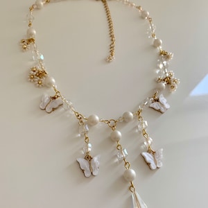 K&ouml;nnte beinhalten: Eine zarte Halskette aus wei&szlig;en Perlen und Kristallen mit einer goldenen Kette und baumelnden Schmetterlingsanh&auml;ngern. Die Halskette hat einen tropfenf&ouml;rmigen Kristallanh&auml;nger.