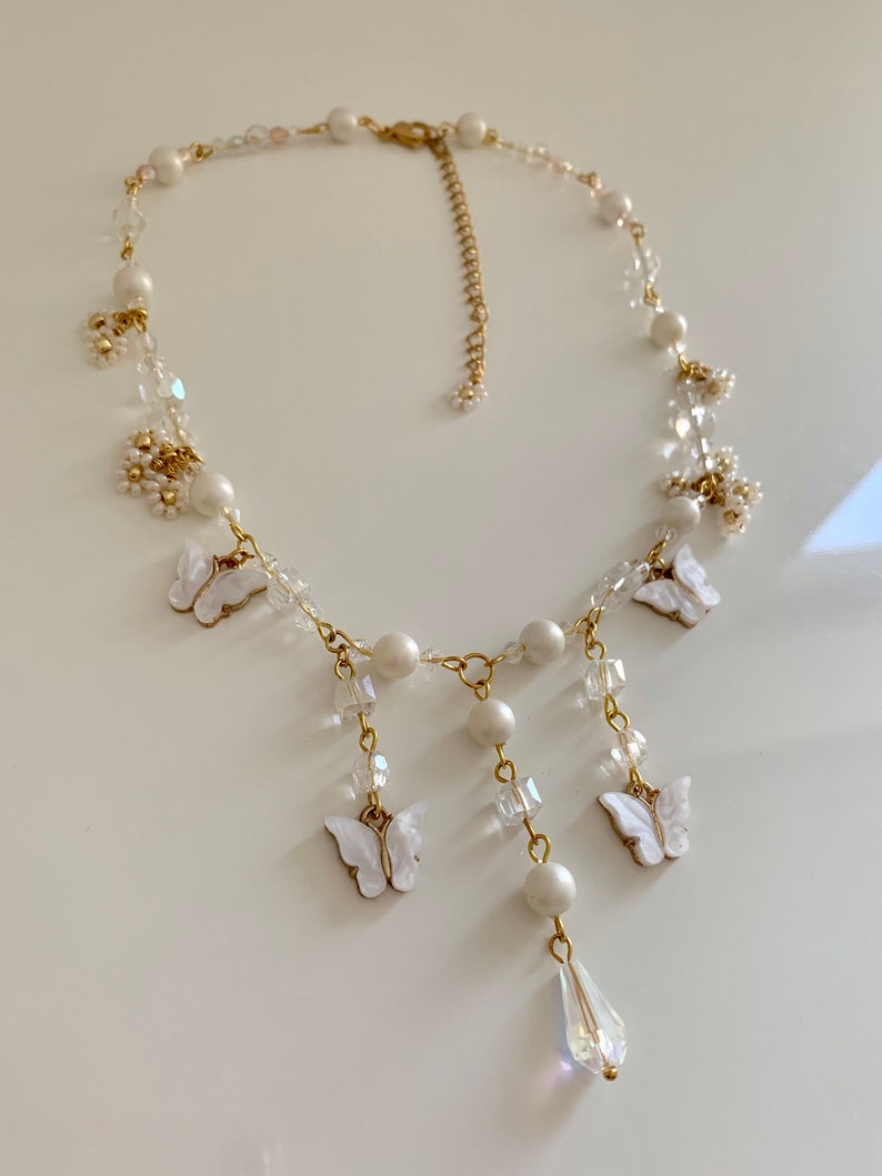 K&ouml;nnte beinhalten: Eine zarte Halskette aus wei&szlig;en Perlen und Kristallen mit einer goldenen Kette und baumelnden Schmetterlingsanh&auml;ngern. Die Halskette hat einen tropfenf&ouml;rmigen Kristallanh&auml;nger.