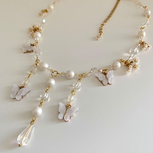 K&ouml;nnte beinhalten: Eine zarte Halskette aus wei&szlig;en Perlen und Kristallen mit einer goldenen Kette und baumelnden Schmetterlingsanh&auml;ngern. Die Halskette hat einen tropfenf&ouml;rmigen Kristallanh&auml;nger.