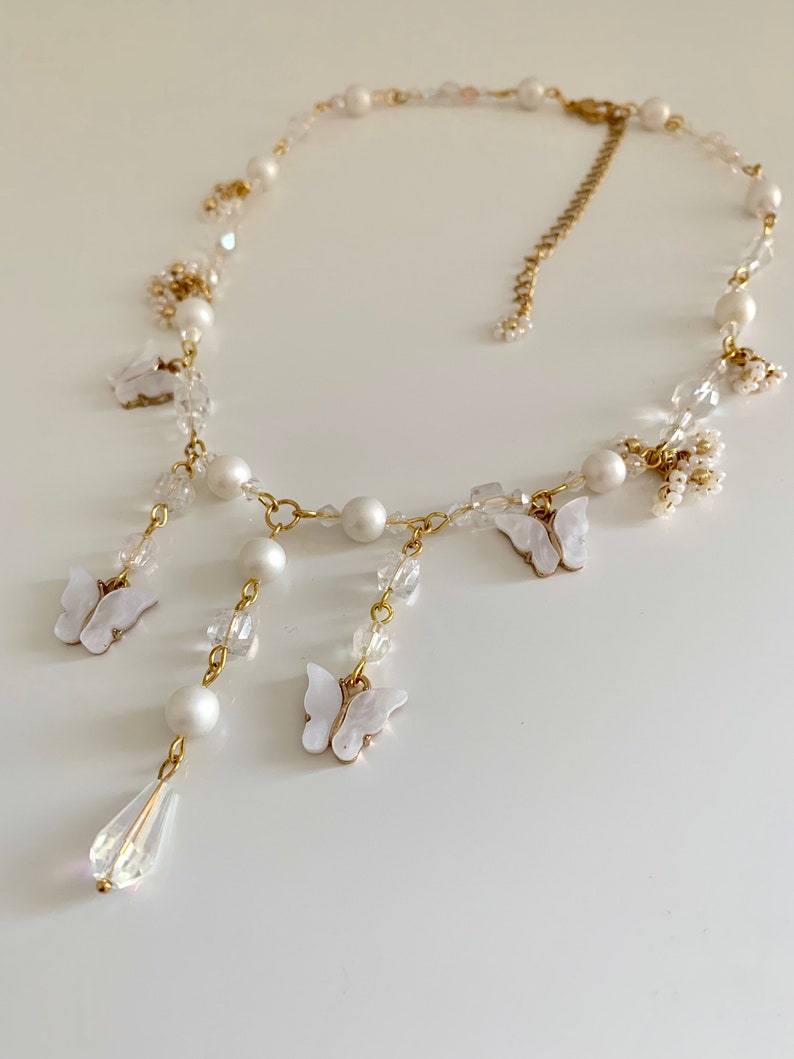 K&ouml;nnte beinhalten: Eine zarte Halskette aus wei&szlig;en Perlen und Kristallen mit einer goldenen Kette und baumelnden Schmetterlingsanh&auml;ngern. Die Halskette hat einen tropfenf&ouml;rmigen Kristallanh&auml;nger.