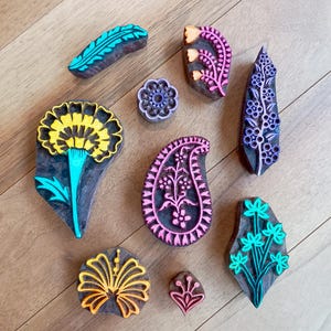 May include: A collection of nine wooden printing blocks featuring various carved designs including a yellow flower, a pink paisley, a purple floral vine, a teal leaf, a teal floral spray, a yellow flower, a small pink flower, and a purple circular motif.