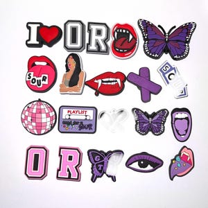 May include: A selection of vibrant shoe charms featuring diverse designs. Includes a red heart, lips with fangs, butterflies, the letters "OR", a disco ball, a cassette tape with "PLAYLIST", and the text "SOUR".