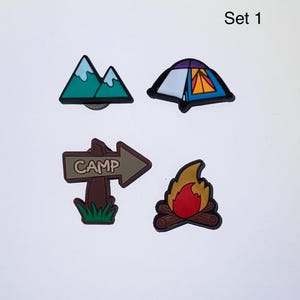 May include: Set of four camping-themed charms. Includes a mountain range with green and white accents, a tent with blue, orange, and purple details, a brown sign with the word "CAMP", and a campfire with red and yellow flames.