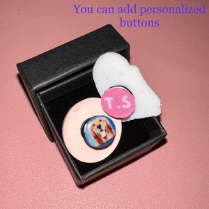 May include: A black gift box contains a light pink phone grip with a photo of a golden retriever, a white decorative piece, and a pink button with the initials "T.S." The text "You can add personalized buttons" is visible.