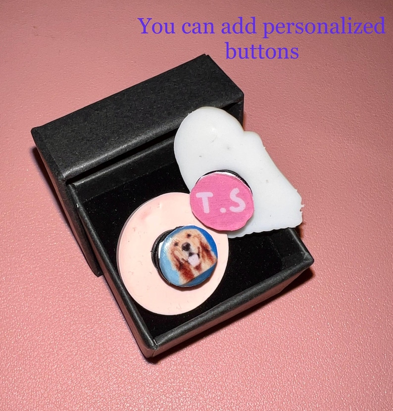 May include: A black gift box contains a light pink phone grip with a photo of a golden retriever, a white decorative piece, and a pink button with the initials "T.S." The text "You can add personalized buttons" is visible.