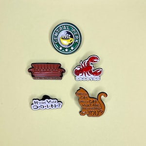May include: A collection of enamel pins featuring designs from the TV show Friends. The pins include the Central Perk logo, a brown couch with "I'll be there for you", a red lobster, a "How You Doin'?" pin, and a brown cat with "Smelly Cat" text.