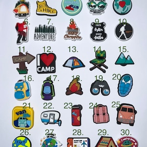 May include: A collection of 30 camping and travel-themed charms. Designs include a tent, hiking boots, a campfire, a backpack, a camper, a globe, and the text "Adventure Awaits". These charms are suitable for shoes, bracelets, or other accessories.