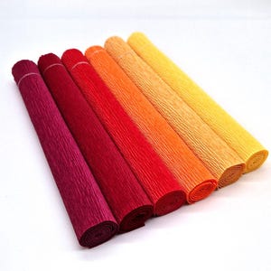 May include: Six rolls of colorful crepe paper in shades of red, orange, and yellow. The paper is tightly rolled and secured with a thin white band. The textured surface of the paper is visible.