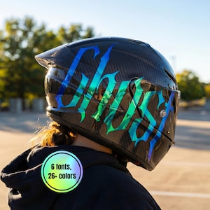 Könnte beinhalten: Schwarzer Motorradhelm mit Karbonfasermuster, mit dem Wort "Ghost" in einer stilisierten, irisierenden Schriftart mit blauen und grünen Farbtönen. Ein runder Aufkleber zeigt "6 Schriftarten, 26+ Farben."