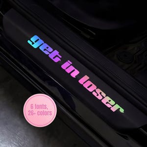 May include: Black car door sill protector with the text "get in loser" in a gradient of blue, purple, and pink. A pink circle with the text "6 fonts, 26+ colours" is in the lower left corner.