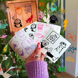 Könnte beinhalten: Eine Sammlung quadratischer, weißer Aufkleberbögen mit verschiedenen schwarzen und rosa grafischen Illustrationen, darunter ein Vampir, ein Totenkopf, ein Kaffeebecher, ein Donut und verschiedene Hunderassen. Der Text JUST A CAT ist aufgedruckt.