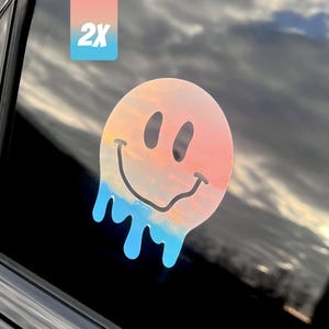 May include: A holographic smiley face sticker with a melting design. The sticker transitions from pink to blue, with a black outline and eyes. A small rectangular label in the upper left corner reads "2X".