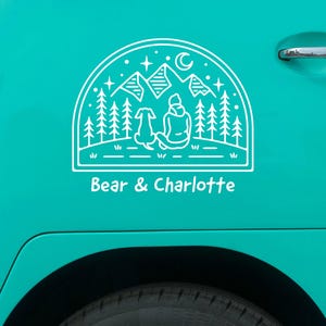 May include: White vinyl decal of a person and a dog sitting in front of a mountain range with a crescent moon and stars. The text "Bear & Charlotte" is below the image.