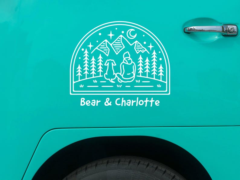 May include: White vinyl decal of a person and a dog sitting in front of a mountain range with a crescent moon and stars. The text "Bear & Charlotte" is below the image.