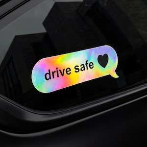 May include: A colorful car decal in the shape of a chat bubble. The bubble has a tie-dye effect with the words "drive safe" in black text and a black heart symbol. The decal is adhered to a car window.