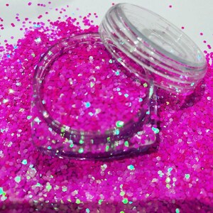 Puede incluir: Un pequeño frasco transparente lleno de purpurina rosa iridiscente. La purpurina está suelta y esparcida alrededor del frasco.