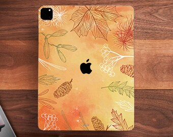 May include: An Apple iPad Pro with a fall-themed skin featuring orange, yellow, and brown leaves, pinecones, and berries. The skin has a watercolor effect and a black Apple logo in the center. The iPad is on a wooden surface.