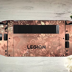 Peut inclure: Une peau décorative pour un appareil de jeu Lenovo Legion. La peau présente un motif floral dans les tons marron et or rose, avec le mot "LEGION" et le logo Lenovo visibles. La peau recouvre l'arrière de l'appareil.