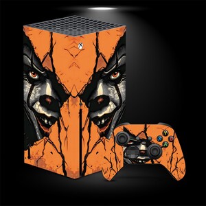 May include: A gaming console and matching wireless controller featuring a graphic skin with an orange, cracked surface design. The artwork displays a clown face with intense eyes and a wide grin, integrated into the fractured orange pattern.